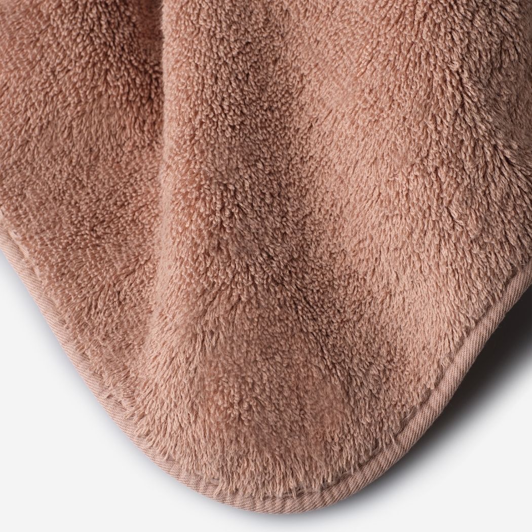 Close-up of a dusty rose cotton towel highlighting the soft terry texture, rounded corner finish, and subtle embroidered logo detail.