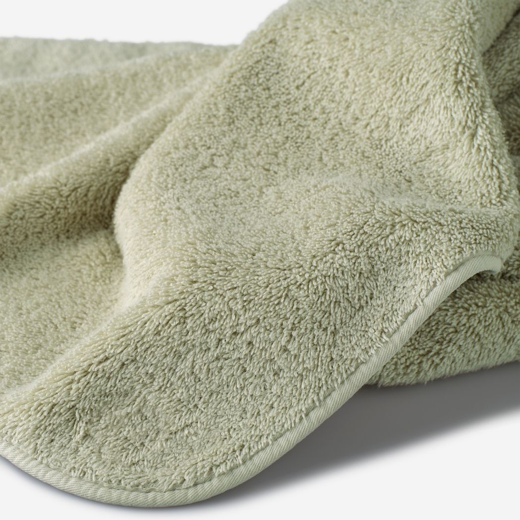 Close-up of a sage green cotton towel highlighting the soft terry texture, rounded corner finish, and subtle embroidered logo detail.