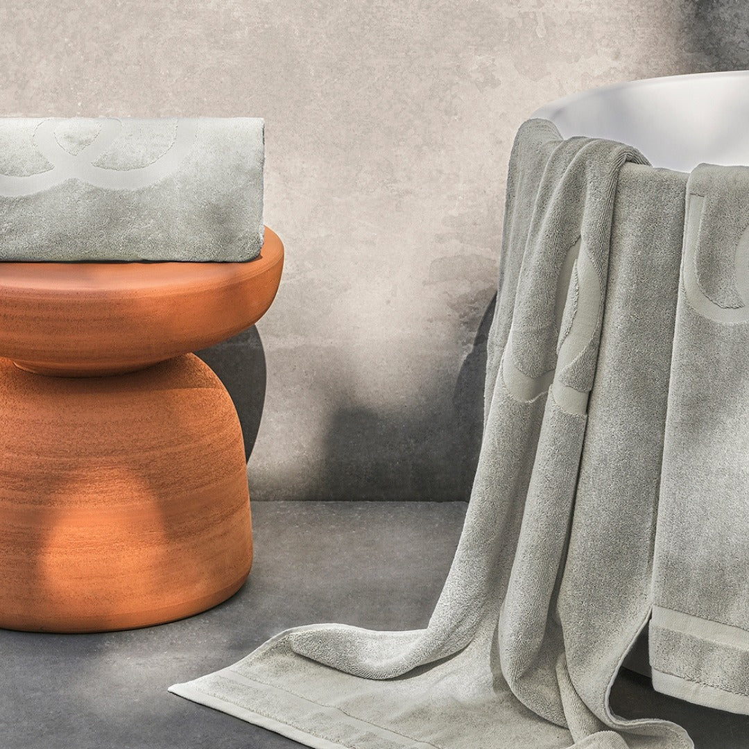 Light grey bath towels with Jacquard logo displayed beside a white bathtub and terracotta stool