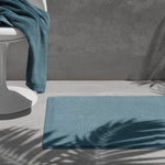 Teal bath mat on a grey floor with a matching towel over a white chair