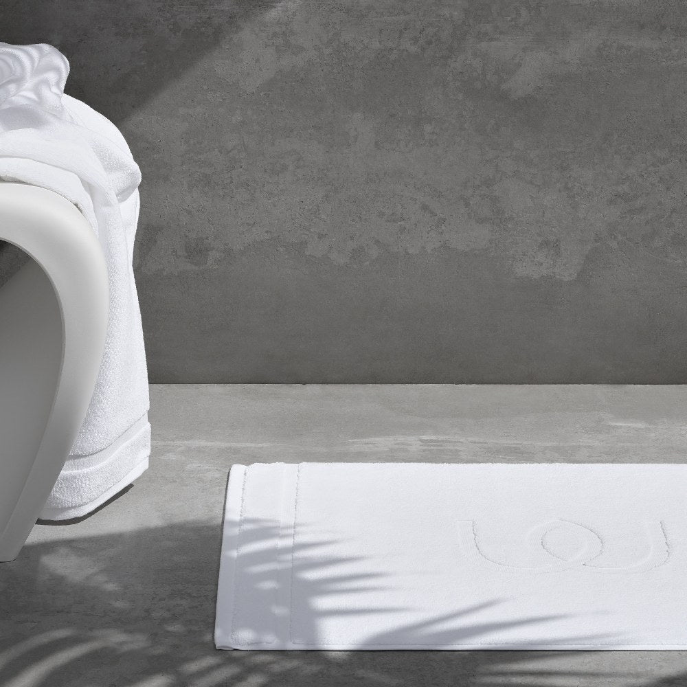 White bath mat on a gray bathroom floor with a textured wall in the background