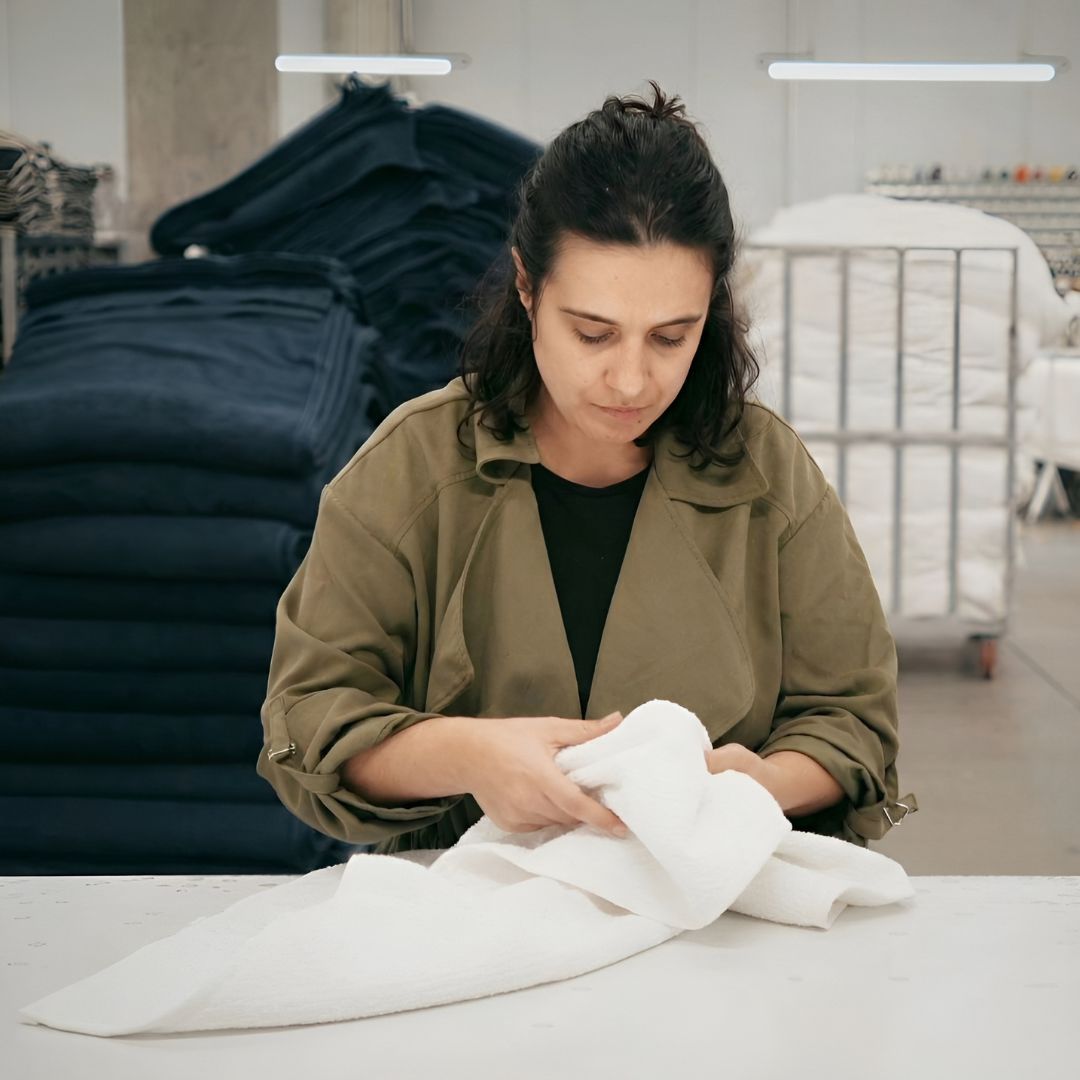 Woman inspecting a white towel in a textile facility with stacks of dark blue fabrics in the background.
